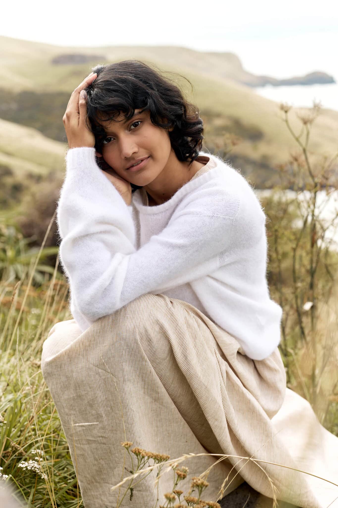 Woman in white sweater crouches in long grass.