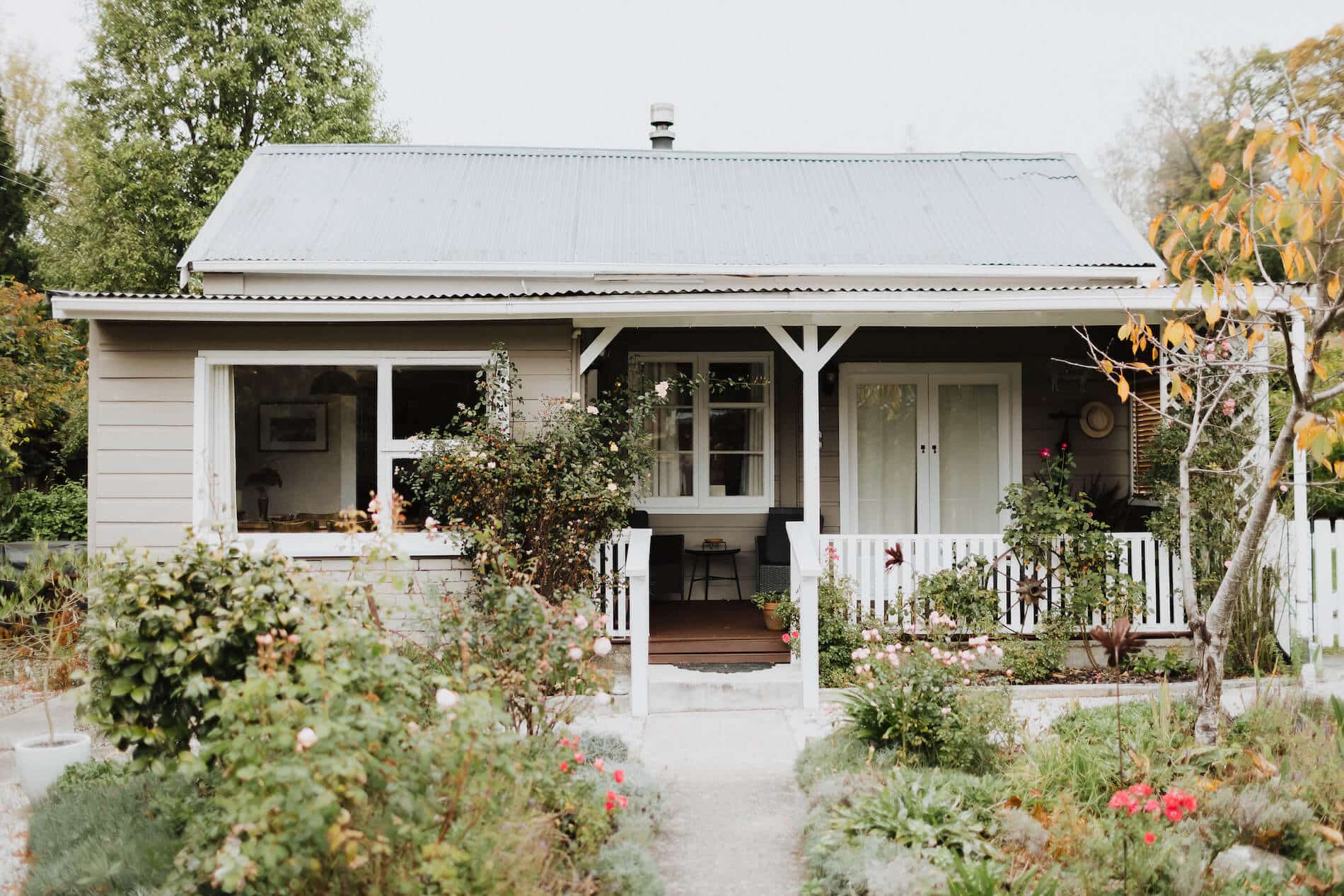 Exterior of a neat/trim cottage from the late 1800s, green and colourful front garden and white covered veranda