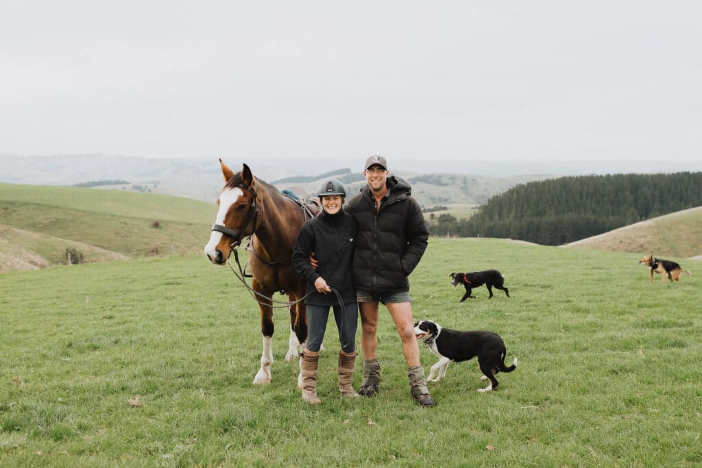 Woman and man stand with their horse and two farm dogs on a big grassy hill