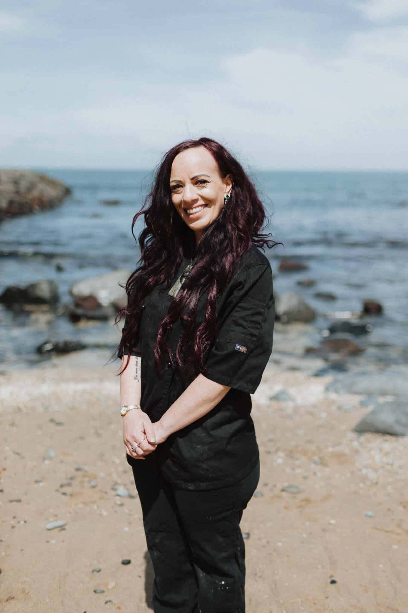 Woman standing on a sandy beach with rocks and ocean in the background.