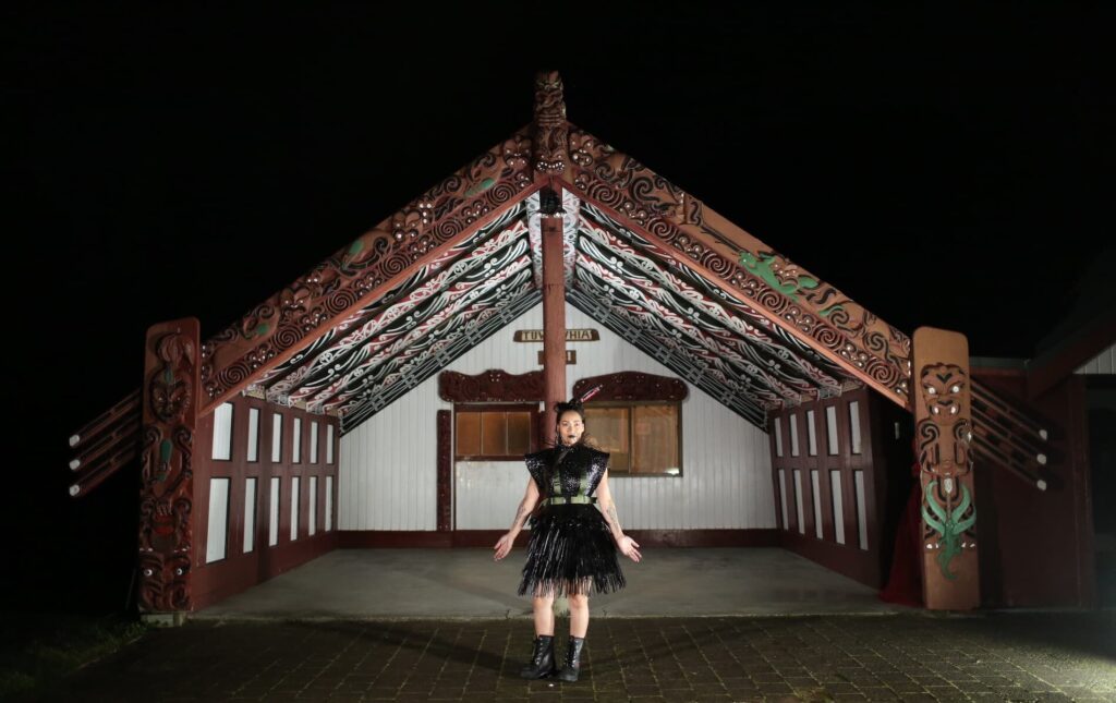 Woman stands in front of marae wearing mahi raranga