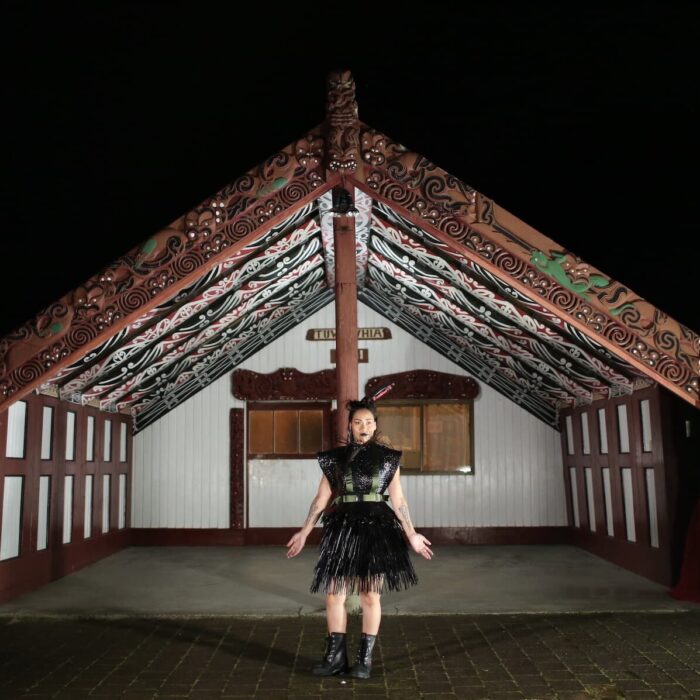 Woman stands in front of marae wearing mahi raranga