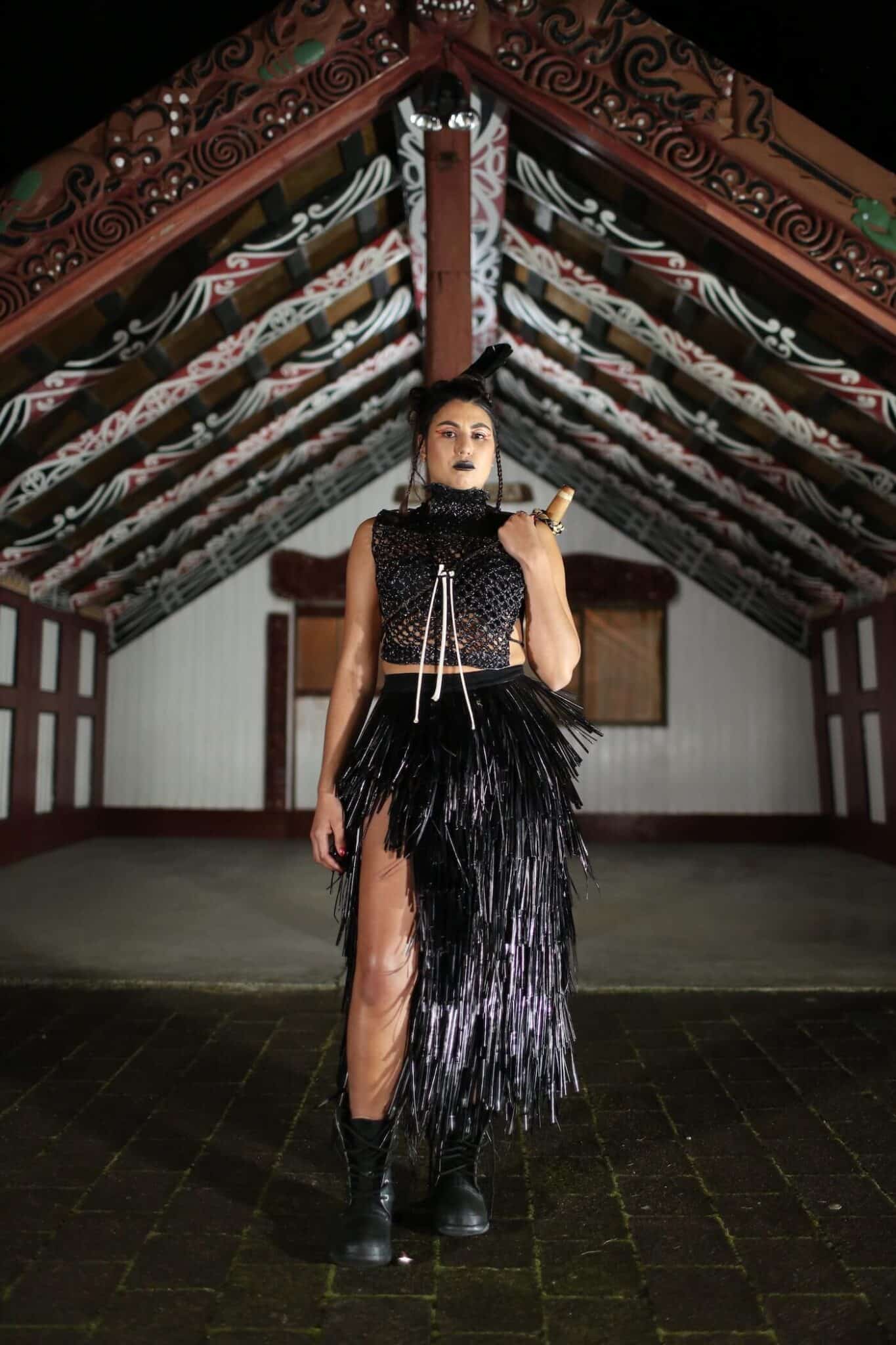 Woman modelling a black mahi raranga in front of a marae