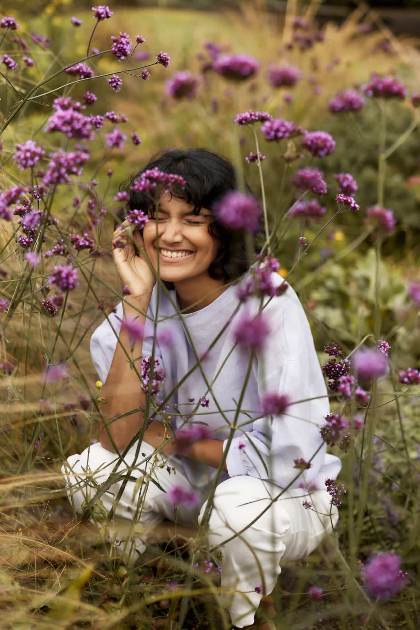 Woman grinning with her eyes closed in the background, out of focus purple flowers in the foreground.
