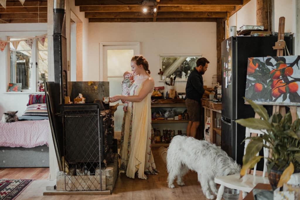 Woman holding her newborn baby stands in the kitchen of her yurt while her husband does something in the background.