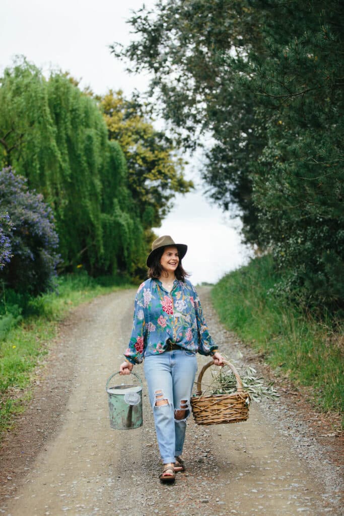 Woman walks down a dirt road holding a watering can and a woven basket of foliage cuttings