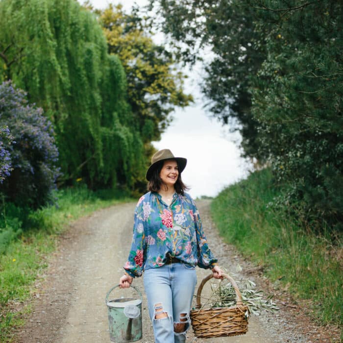 Woman walks down a dirt road holding a watering can and a woven basket of foliage cuttings
