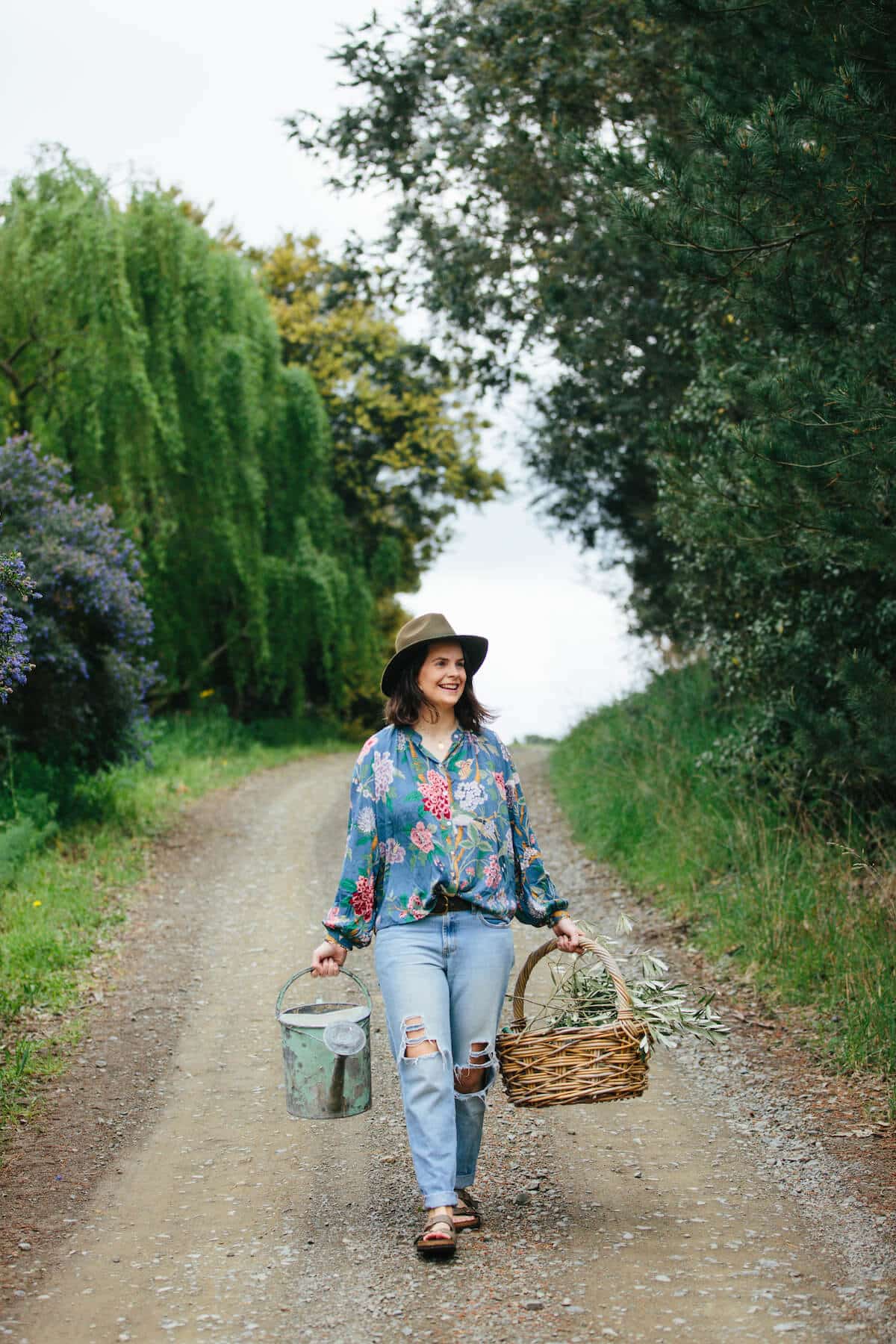 Woman walks down a dirt road holding a watering can and a woven basket of foliage cuttings