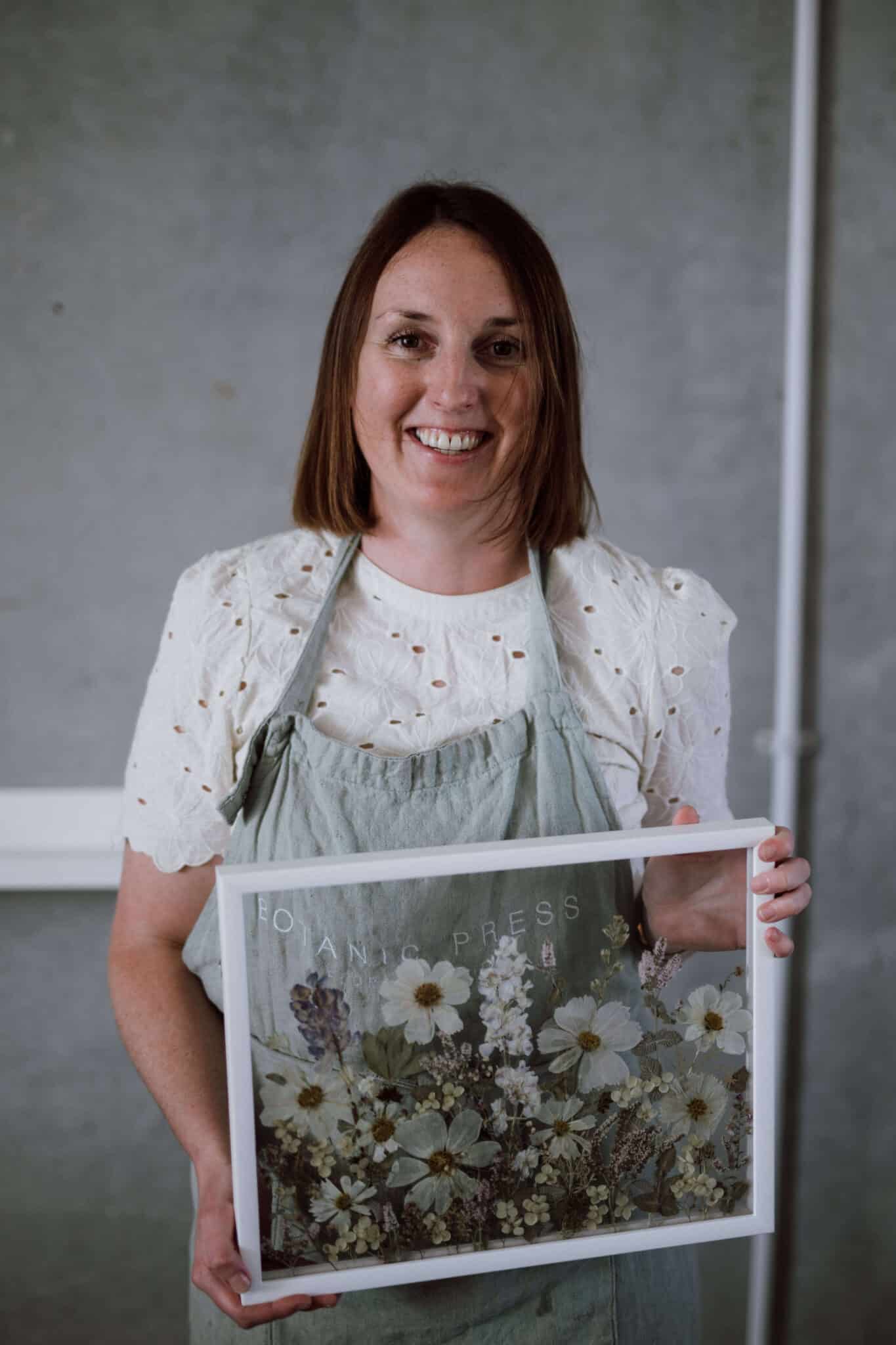 Woman smiles as she holds a framed glass pressed floral artwork.