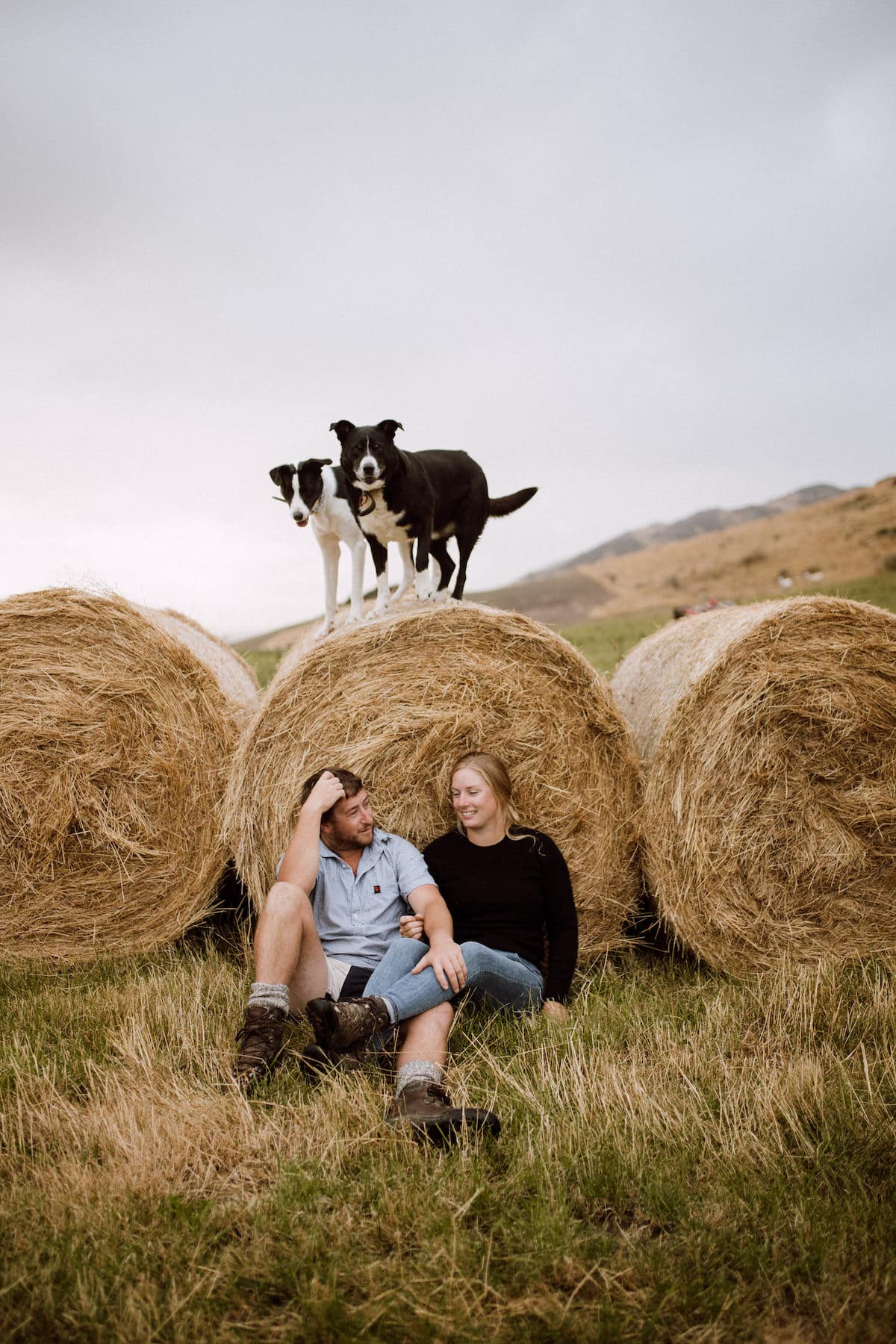 Ilka, who is German, and her Kiwi partner Callum with their two dogs sit in front of hay bales on the farm.