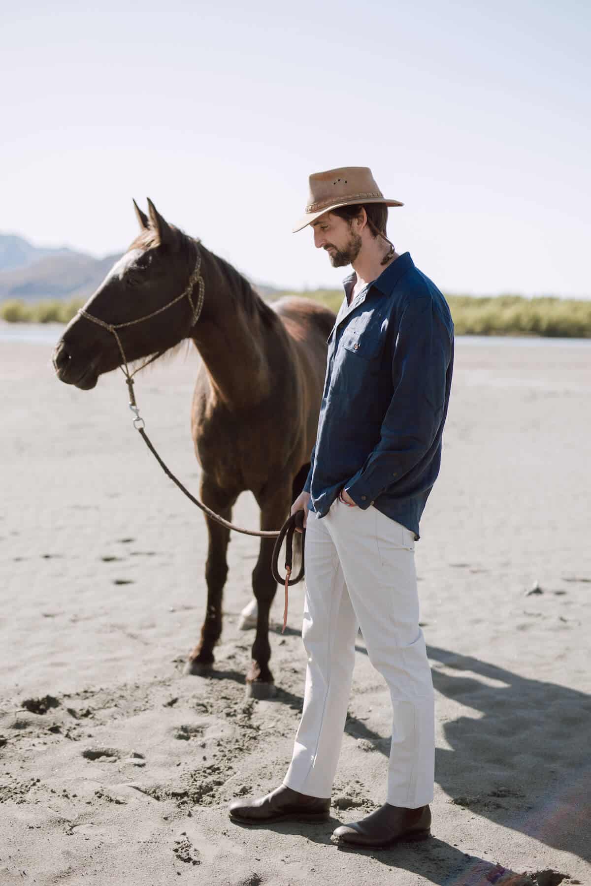 Man holds a horse by the reins on a sandy piece of land.