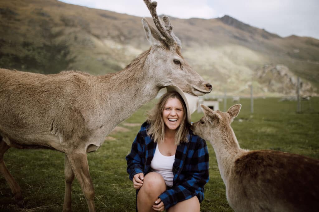 Women kneeling between two deer laughs as they knock her hat off.