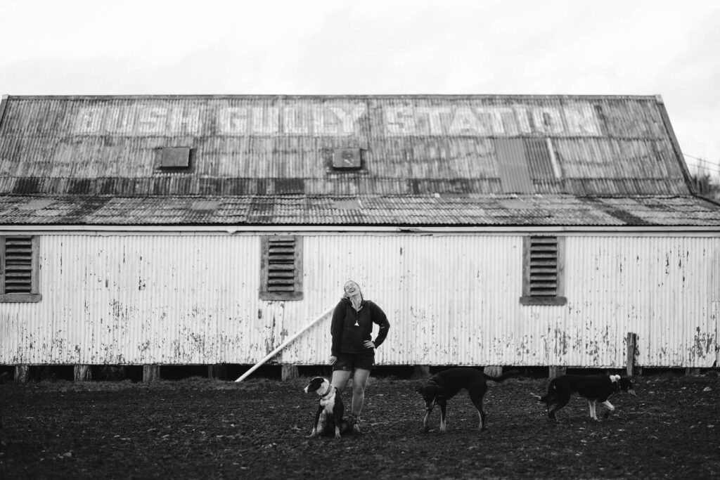 Black and white image of a young female shepherd standing in front of a large barn with two dogs.
