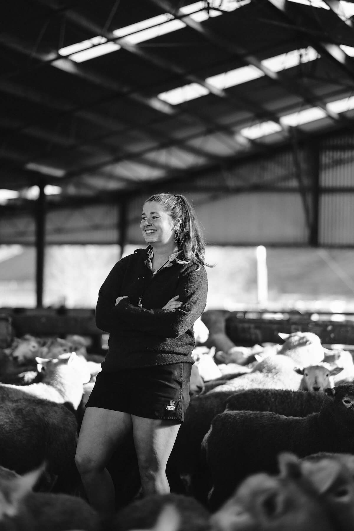 Black and white image of a young female shepherd standing amongst sheep in a big shed.