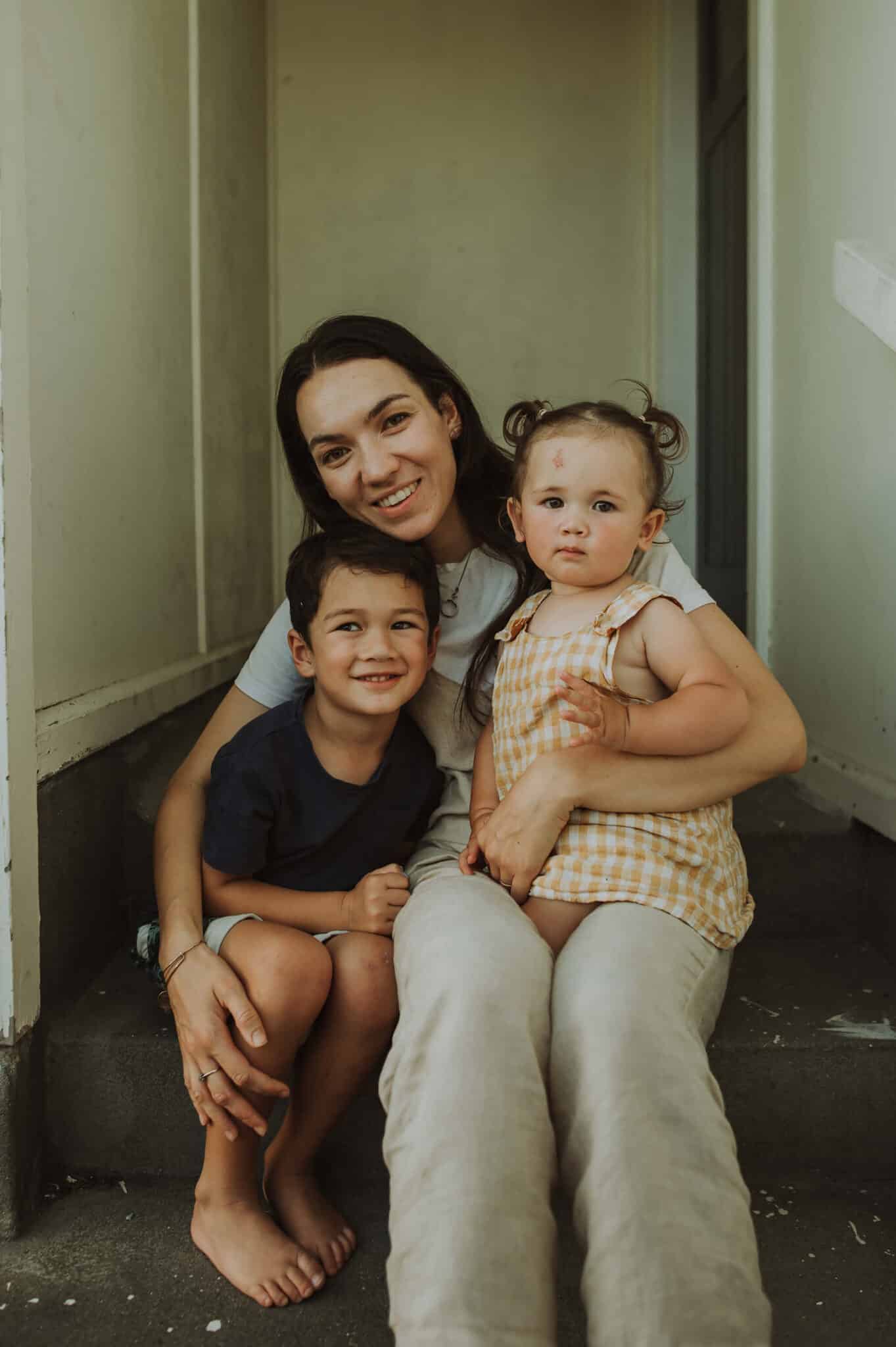 Woman and her two young children smile at the camera, sitting on a step