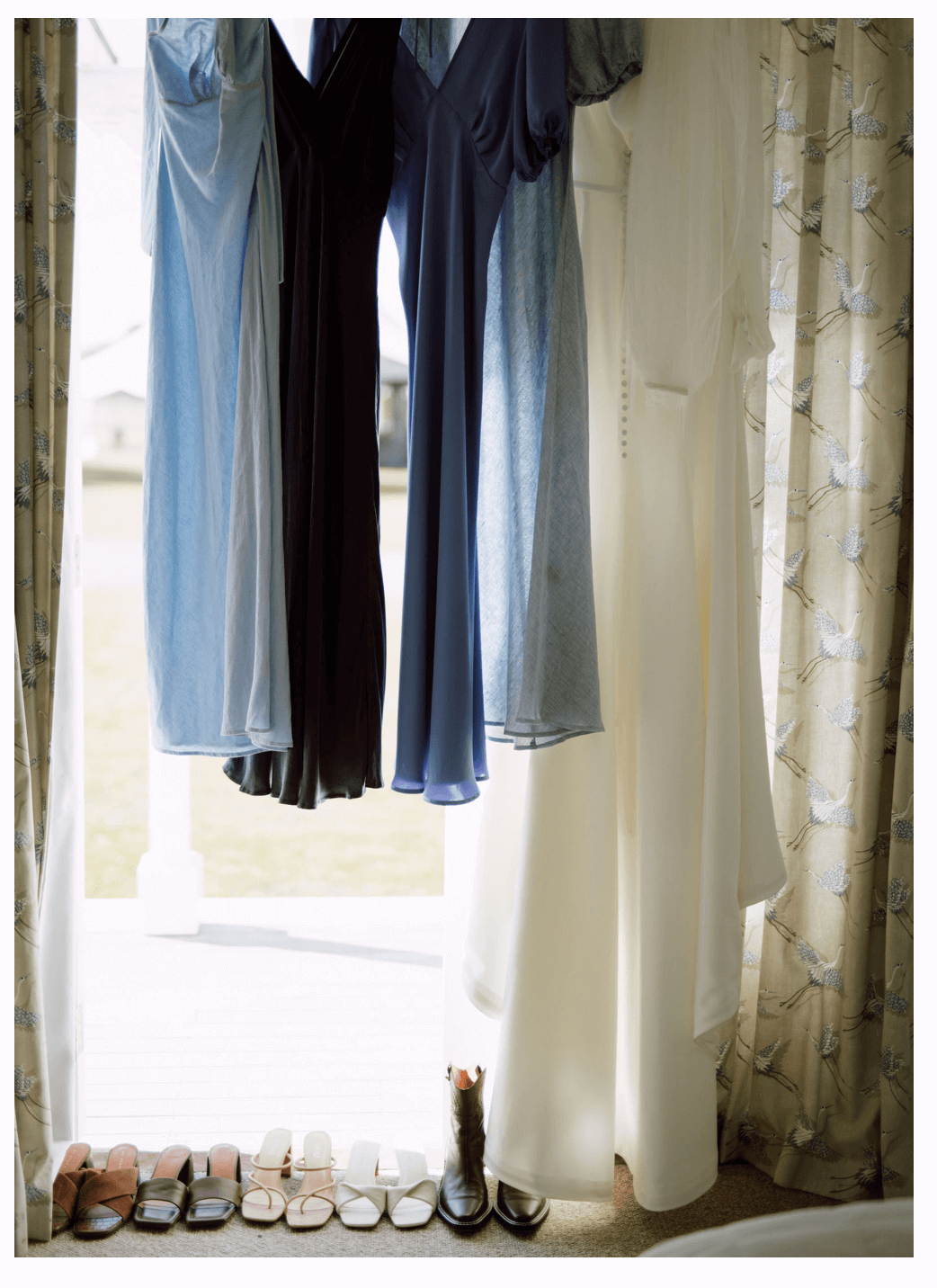 Blue bridesmaids' dresses and white bride's dress hang in front of a window.