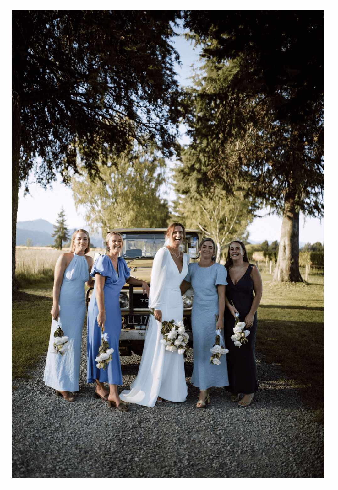 Bridal party smiles for a photo against a vintage Toyota Land Cruiser on a tree-lined driveway.