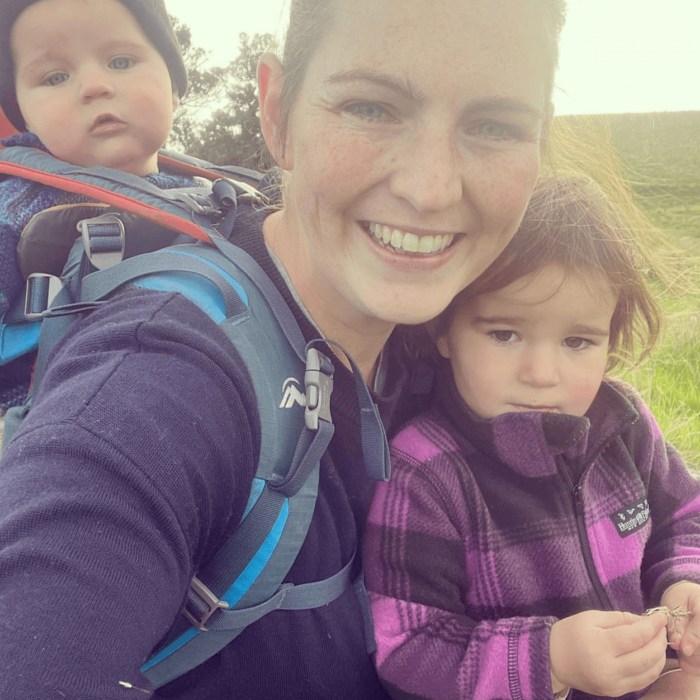 Woman smiles as she takes a photo of herself with one child in a baby backpack on her back and another held on her front.