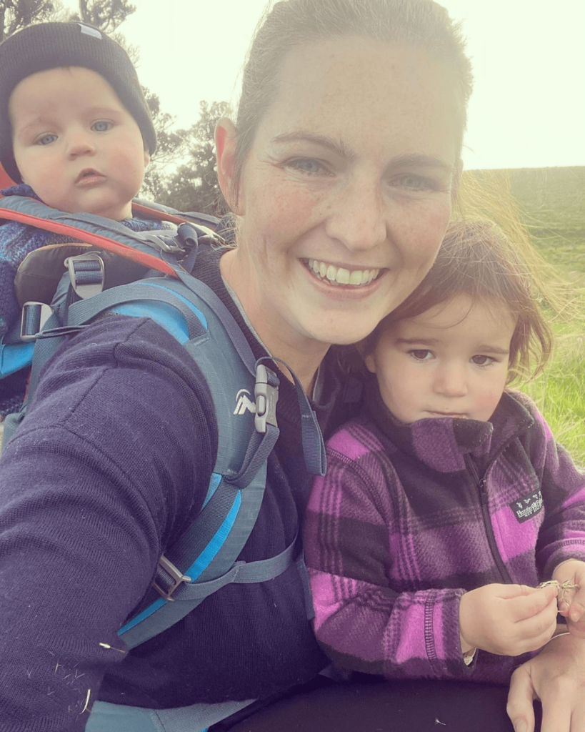 Woman smiles as she takes a photo of herself with one child in a baby backpack on her back and another held on her front.