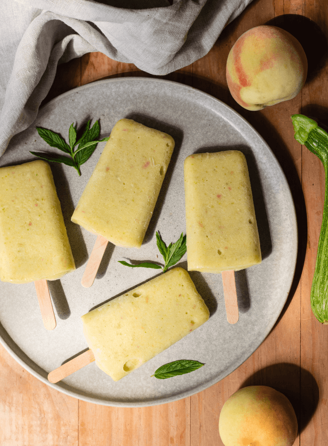 Four cream coloured ice lollies sit on a plate surrounded by sprigs of mint, peaches and a zucchini