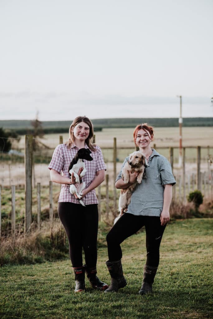 Two women stand in a paddock, holding a puppy each.
