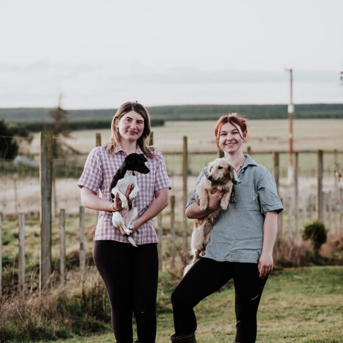 Two women stand in a paddock, holding a puppy each.