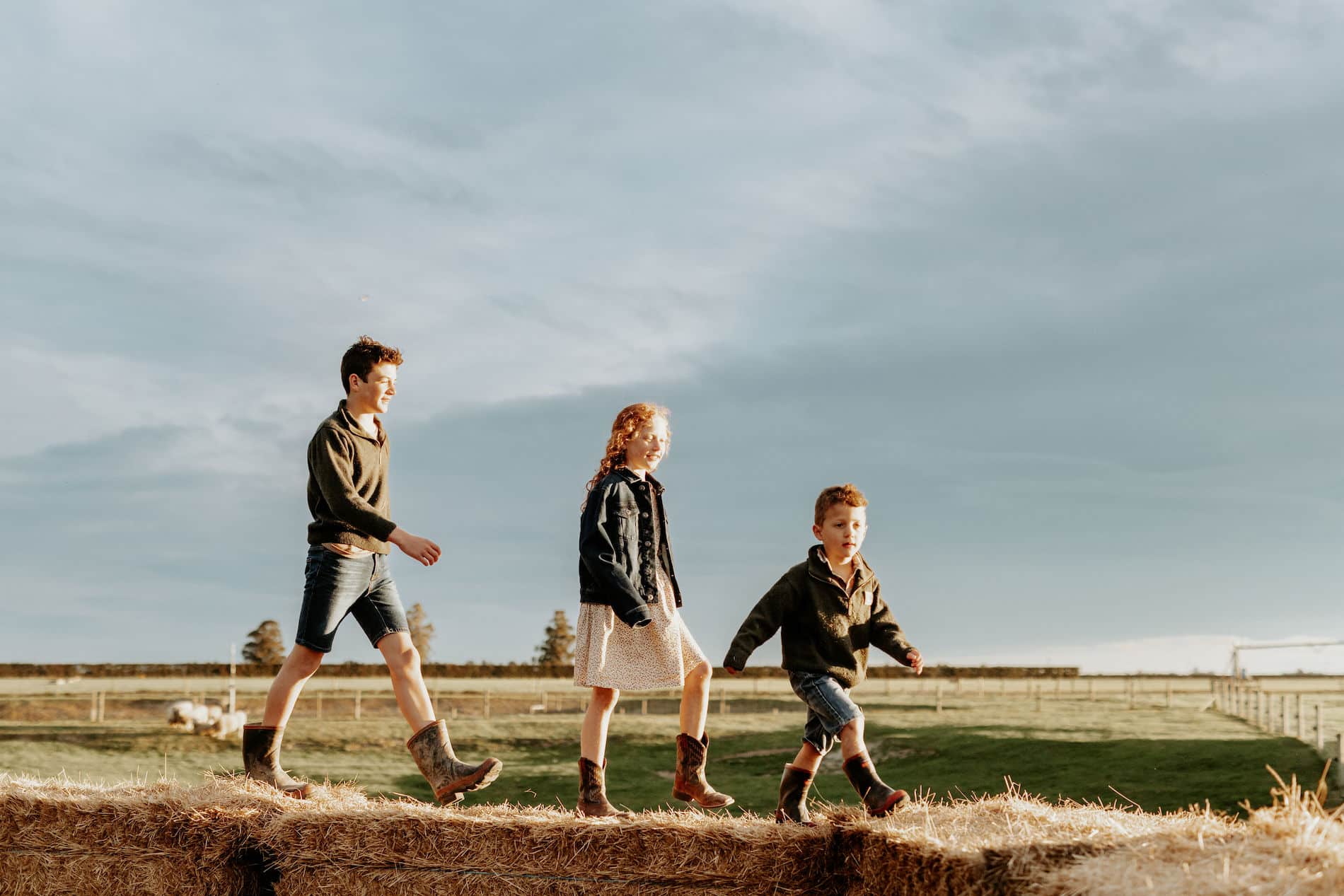Three kids walk along hay bales on a farm.