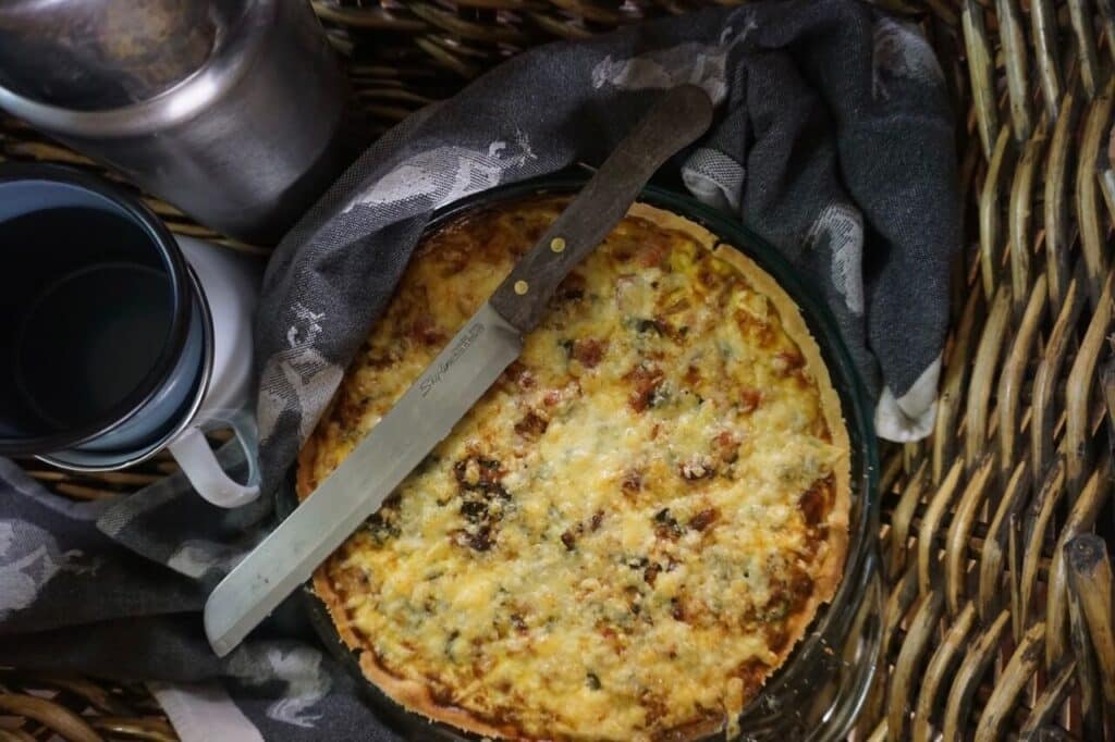 Quiche sits in a basket, wrapped in a dish towel and with a knife on top.