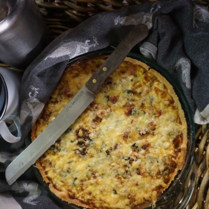 Quiche sits in a basket, wrapped in a dish towel and with a knife on top.