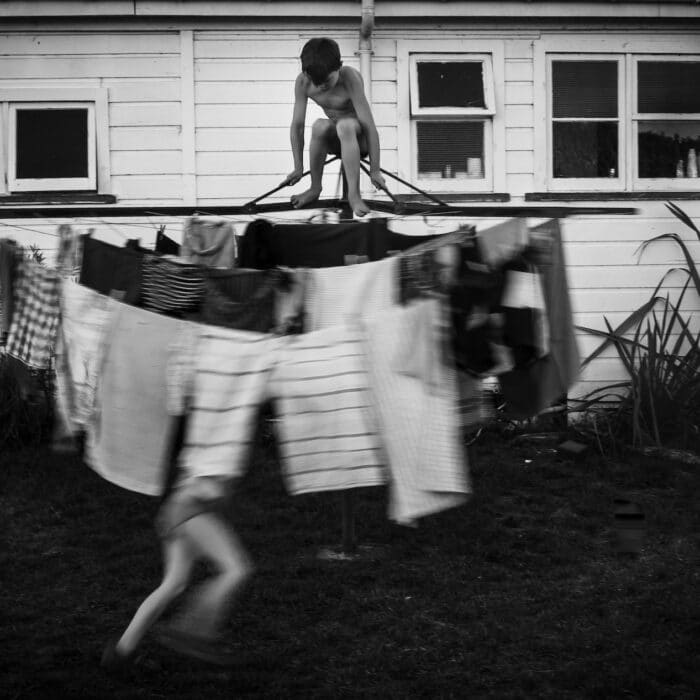 Black and white image of a shirtless boy on top of a washing line that is being spun around by his sister, blurred, in motion