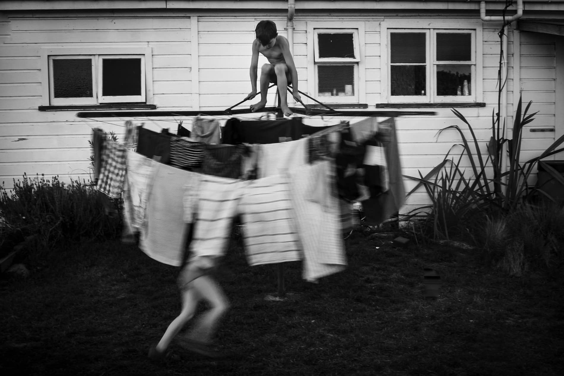 Black and white image of a shirtless boy on top of a washing line that is being spun around by his sister, blurred, in motion
